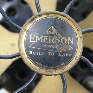 Puede incluir: Primer plano de un ventilador Emerson antiguo. Las aspas del ventilador son doradas y la carcasa del motor es negra con un emblema dorado que dice "Fans Emerson Motors Built to Last."