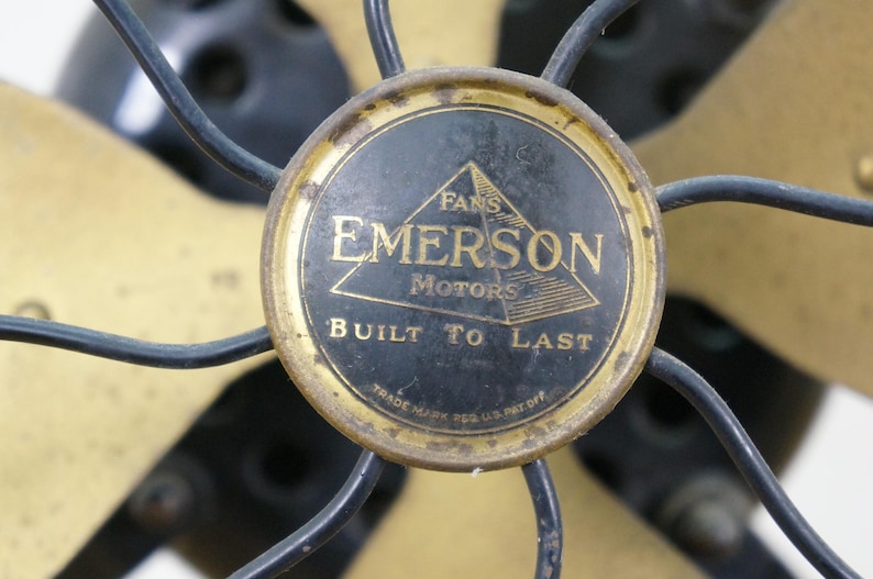 Puede incluir: Primer plano de un ventilador Emerson antiguo. Las aspas del ventilador son doradas y la carcasa del motor es negra con un emblema dorado que dice "Fans Emerson Motors Built to Last."