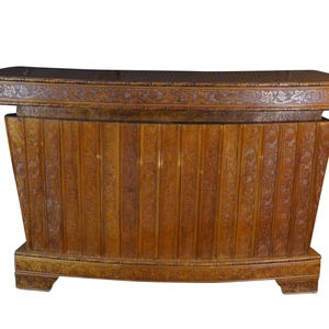 May include: A rectangular wooden bar counter with a polished amber finish. The front features vertical panels with intricate carved floral and vine motifs. The top edge is decorated with a horizontal band of inlaid silver-toned leaf patterns.