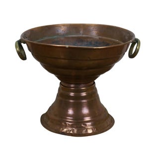 May include: A vintage copper bowl with a pedestal base and two brass handles. The bowl has a wide, open top and a tiered design. The copper has a rich, aged patina, giving it a rustic appearance. The bowl is likely used for decorative purposes.