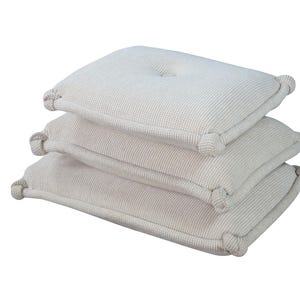 May include: A stack of three rectangular off-white cushions featuring a textured waffle-knit fabric. Each cushion has a central tufted button detail and thick, knotted corner accents that provide a distinct border to the soft, padded structure.