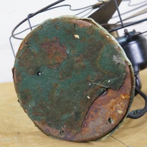 Puede incluir: Primer plano de un ventilador el&eacute;ctrico antiguo. La base es redonda con una cubierta de fieltro verde, que muestra signos de desgaste y &oacute;xido. La carcasa del motor del ventilador es negra y las aspas son visibles en el fondo.