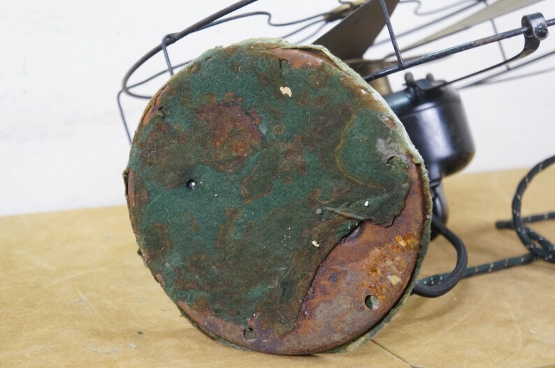 Puede incluir: Primer plano de un ventilador el&eacute;ctrico antiguo. La base es redonda con una cubierta de fieltro verde, que muestra signos de desgaste y &oacute;xido. La carcasa del motor del ventilador es negra y las aspas son visibles en el fondo.