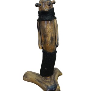 May include: A handcrafted wooden figurine with a stylized humanoid form. The figure has a rounded head with small, dark protrusions and is adorned with a black fabric collar and lower garment. The base is a naturalistic, branch-like shape.