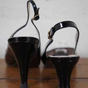 May include: A pair of black patent leather heeled slingback shoes with gold buckles. The shoes are on a wooden surface.