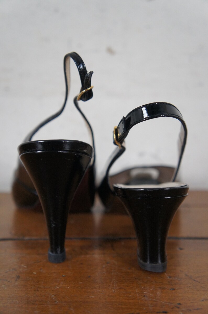 May include: A pair of black patent leather heeled slingback shoes with gold buckles. The shoes are on a wooden surface.