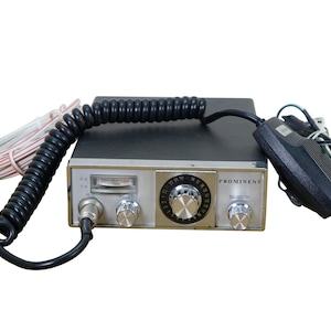 May include: Vintage CB radio with a black and silver design. The radio features a prominent dial, multiple knobs, and a microphone. The text "PROMINENT" is visible on the face of the radio.