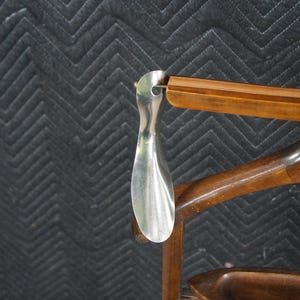 May include: A silver shoe horn hangs from a wooden chair. The shoe horn is a long, curved piece of metal. The chair is made of dark wood and has a curved back and a small tray.