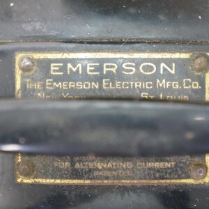 Puede incluir: Primer plano de un motor el&eacute;ctrico Emerson vintage. La carcasa de metal negro presenta dos placas de lat&oacute;n rectangulares con texto en relieve. La placa superior dice "EMERSON" y "The Emerson Electric Mfg. Co."