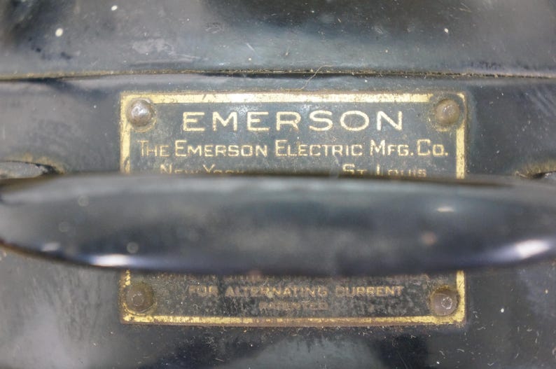 Puede incluir: Primer plano de un motor el&eacute;ctrico Emerson vintage. La carcasa de metal negro presenta dos placas de lat&oacute;n rectangulares con texto en relieve. La placa superior dice "EMERSON" y "The Emerson Electric Mfg. Co."