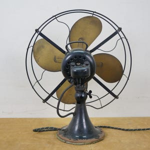 Puede incluir: Un ventilador el&eacute;ctrico antiguo con marco y base de metal negro. Las aspas del ventilador son de color dorado y la jaula es de alambre negro. El ventilador tiene una carcasa de motor verde oscuro y un cable de alimentaci&oacute;n negro y verde. El ventilador est&aacute; sobre una superficie de madera.