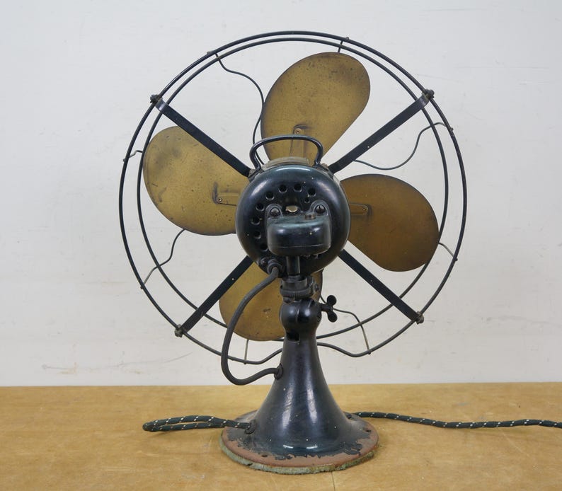 Puede incluir: Un ventilador el&eacute;ctrico antiguo con marco y base de metal negro. Las aspas del ventilador son de color dorado y la jaula es de alambre negro. El ventilador tiene una carcasa de motor verde oscuro y un cable de alimentaci&oacute;n negro y verde. El ventilador est&aacute; sobre una superficie de madera.