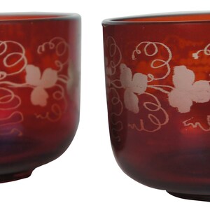 May include: Two red glass cups with a white grapevine design. The cups are clear and have a slightly rounded shape.