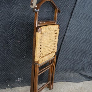 May include: A vintage wooden valet chair with a woven seat and a coat hanger. The chair is made of dark wood and features a metal shoehorn. The seat is woven with a light brown rope. The chair is folded.