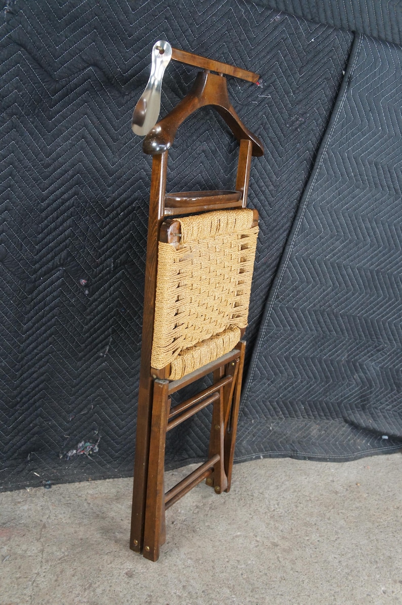 May include: A vintage wooden valet chair with a woven seat and a coat hanger. The chair is made of dark wood and features a metal shoehorn. The seat is woven with a light brown rope. The chair is folded.