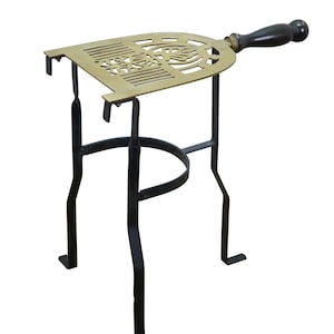 May include: A brass trivet with a decorative cutout pattern and a dark wooden handle, supported by a black metal stand with three legs. The stand features a circular ring connecting the legs, providing a stable base for the elevated brass top.