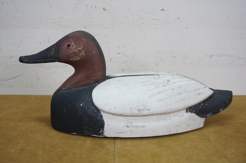 May include: A wooden duck decoy, painted in shades of white, black, and brown. The duck has a brown head and a black beak. The body is primarily white with black accents. The decoy is designed to float on water.