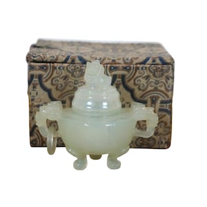 May include: A pale green jade incense burner with dragon-shaped handles and a decorative lid. The burner sits in front of a patterned box with a beige and blue design. The incense burner is a small, detailed decorative item.