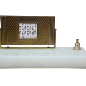 May include: A vintage desk calendar with a brass frame and a white marble base. The calendar displays the days of the month. A small brass object sits on the right side of the base. The calendar is 4 inches wide.