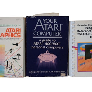 May include: Three vintage Atari computer books with colorful covers. The first book is titled "Computers First Book of Atari Graphics". The second book is titled "Your Atari Computer: A Guide to Atari 400/800 Personal Computers". The third book is titled "Computer Direct Programmer's Reference Guide for the Atari 400/800 Computers".