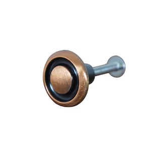 May include: A round, copper-colored cabinet knob with a black ring around the edge and a smaller copper-colored circle in the center. The knob has a silver screw attached to the back.