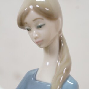 May include: A porcelain figurine of a woman with long blonde hair in a ponytail, wearing a light blue dress. The figurine has a serene expression, with her head tilted downward. The details are finely crafted, showcasing a delicate and elegant design.