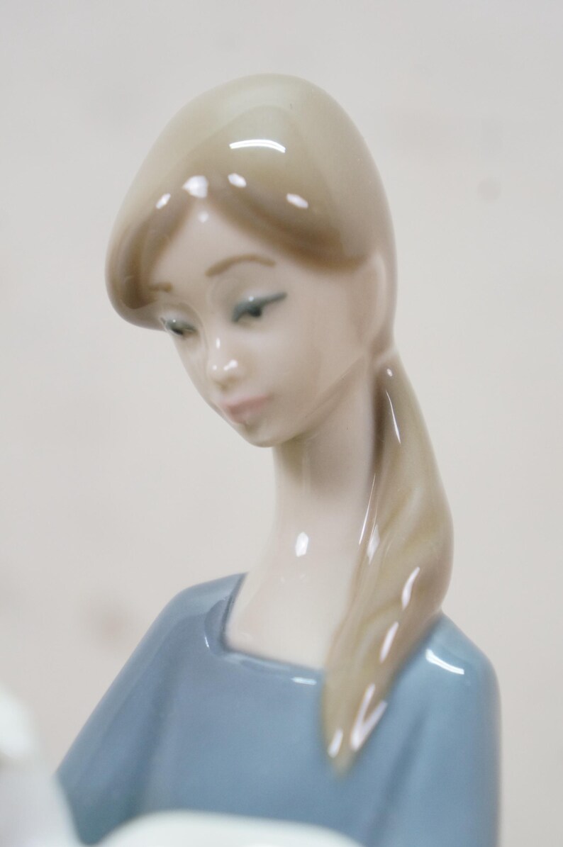 May include: A porcelain figurine of a woman with long blonde hair in a ponytail, wearing a light blue dress. The figurine has a serene expression, with her head tilted downward. The details are finely crafted, showcasing a delicate and elegant design.