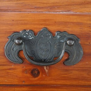Pode incluir: Close-up de um puxador de gaveta de metal escuro com um design decorativo, montado em uma superf&iacute;cie de madeira. O puxador tem uma al&ccedil;a curva e detalhes ornamentados, com um acabamento escuro. A madeira tem um tom quente, marrom avermelhado.