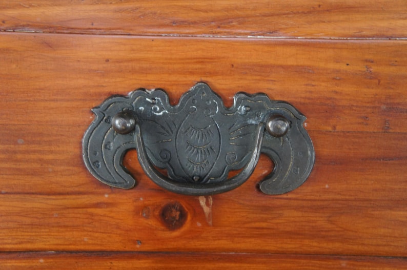 Pode incluir: Close-up de um puxador de gaveta de metal escuro com um design decorativo, montado em uma superf&iacute;cie de madeira. O puxador tem uma al&ccedil;a curva e detalhes ornamentados, com um acabamento escuro. A madeira tem um tom quente, marrom avermelhado.