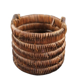 May include: A woven brown basket made of natural materials. The basket is cylindrical in shape and has a smooth, even texture.