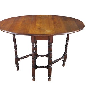 May include: A brown wooden drop-leaf table with an oval top and a smooth, polished surface. The table features four turned legs connected by horizontal stretchers, providing structural support. The wood grain is visible across the tabletop.