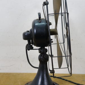Puede incluir: Un ventilador el&eacute;ctrico antiguo con una carcasa de motor negra, aspas de lat&oacute;n y una jaula protectora de alambre. El ventilador tiene una base de pedestal y un cable de alimentaci&oacute;n. El dise&ntilde;o recuerda a los electrodom&eacute;sticos de principios del siglo XX.