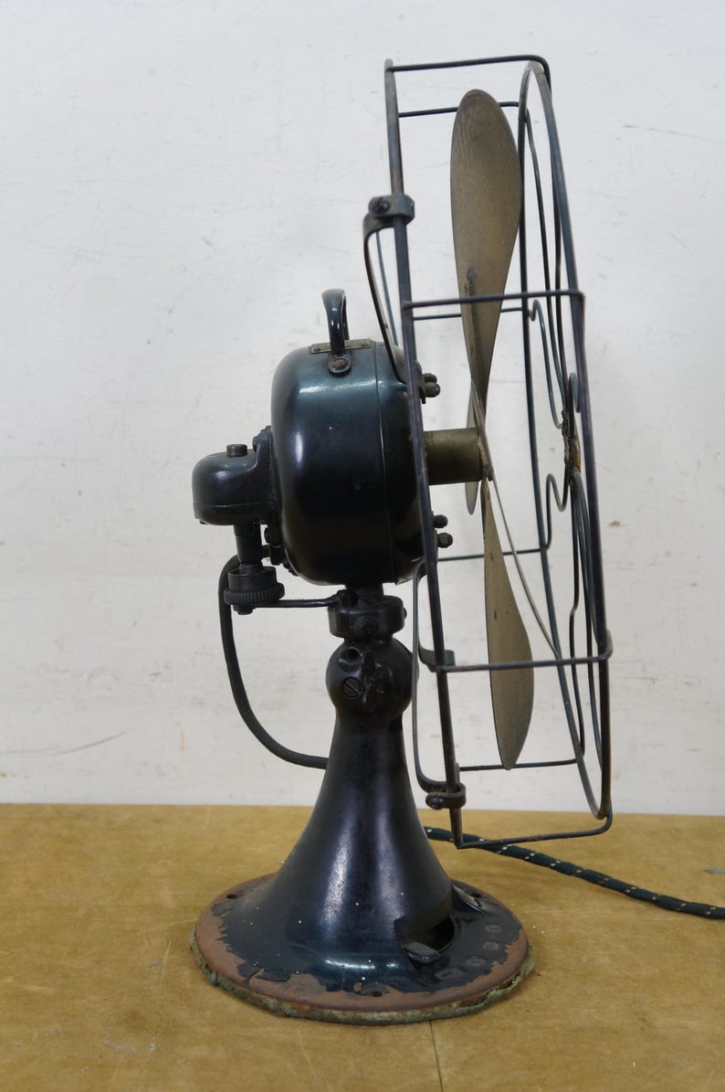 Puede incluir: Un ventilador el&eacute;ctrico antiguo con una carcasa de motor negra, aspas de lat&oacute;n y una jaula protectora de alambre. El ventilador tiene una base de pedestal y un cable de alimentaci&oacute;n. El dise&ntilde;o recuerda a los electrodom&eacute;sticos de principios del siglo XX.
