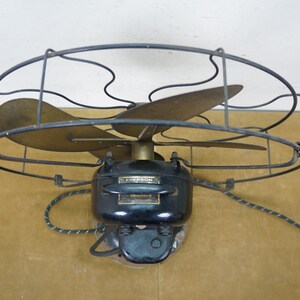 Puede incluir: Ventilador el&eacute;ctrico Emerson vintage con tres aspas de lat&oacute;n y una carcasa de motor negra. El ventilador est&aacute; encerrado en una jaula de alambre y tiene un cable de alimentaci&oacute;n con un patr&oacute;n de rayas.
