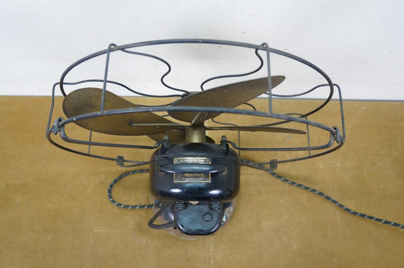 Puede incluir: Ventilador el&eacute;ctrico Emerson vintage con tres aspas de lat&oacute;n y una carcasa de motor negra. El ventilador est&aacute; encerrado en una jaula de alambre y tiene un cable de alimentaci&oacute;n con un patr&oacute;n de rayas.