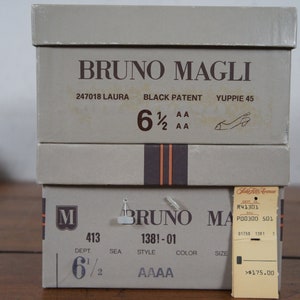 May include: Two shoe boxes stacked on top of each other. The top box is labeled "BRUNO MAGLI" and has the text "247018 LAURA BLACK PATENT YUPPIE 45 6 1/2 AA 2 AA". The bottom box is labeled "BRUNO MAGLI" and has the text "M 413 DEPT 6 1/2 SEA 1381-01 STYLE COLOR SIZE AAAA". A price tag is attached to the bottom box with the text "R41301 P00300 501 01753 1381 1 >$175.00".