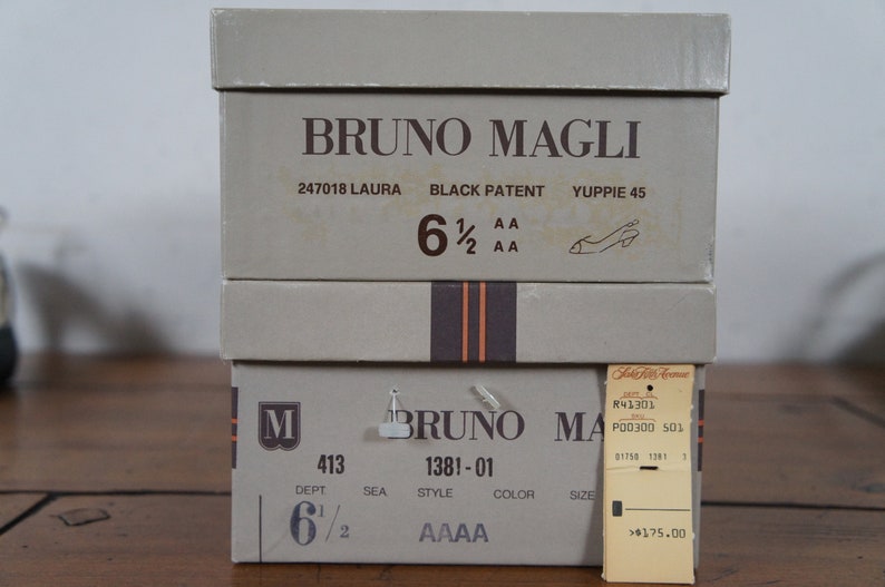 May include: Two shoe boxes stacked on top of each other. The top box is labeled "BRUNO MAGLI" and has the text "247018 LAURA BLACK PATENT YUPPIE 45 6 1/2 AA 2 AA". The bottom box is labeled "BRUNO MAGLI" and has the text "M 413 DEPT 6 1/2 SEA 1381-01 STYLE COLOR SIZE AAAA". A price tag is attached to the bottom box with the text "R41301 P00300 501 01753 1381 1 >$175.00".