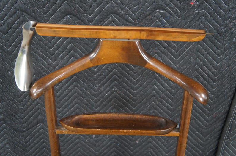 May include: A wooden valet stand with a built-in clothes hanger and a small tray. A silver-colored shoehorn is attached to the side. The stand is made of dark-stained wood and has a classic design.