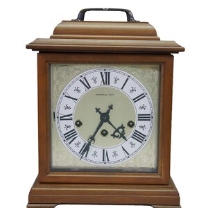 May include: A wooden mantel clock with a rectangular frame and a top handle. The clock face is cream-colored with black Roman numerals, ornate black hands, and the word Hamilton printed in the center. The background features a subtle floral pattern.
