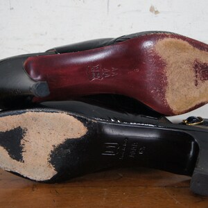 May include: A pair of black leather heeled shoes with a gold buckle. The shoes have a pointed toe and a high heel. The soles of the shoes are visible.