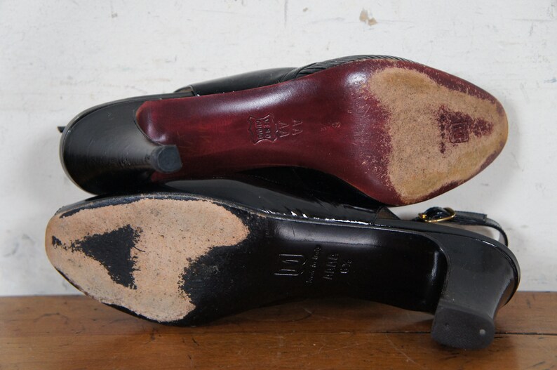 May include: A pair of black leather heeled shoes with a gold buckle. The shoes have a pointed toe and a high heel. The soles of the shoes are visible.