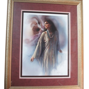 May include: A framed print of a Native American woman in a traditional dress, standing in a soft, ethereal light. The woman is wearing turquoise jewelry and has long, dark hair. The print is signed by the artist.