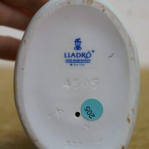 May include: Underside of a white porcelain figurine with the Lladro logo and the text "HAND MADE IN SPAIN". The number "4505" is printed in black, along with a blue sticker with the number "205".