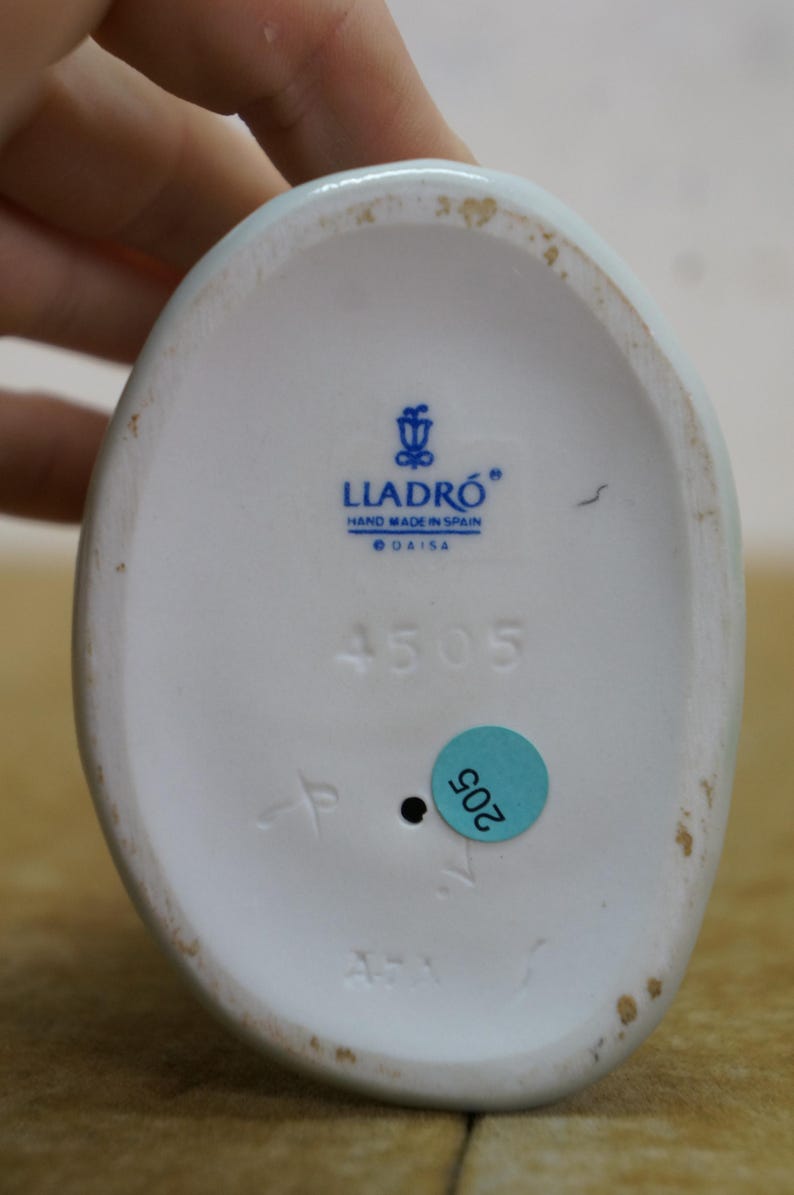 May include: Underside of a white porcelain figurine with the Lladro logo and the text "HAND MADE IN SPAIN". The number "4505" is printed in black, along with a blue sticker with the number "205".