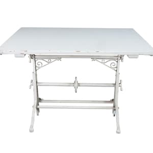 May include: A white drafting table with a rectangular top and a metal base. The base features decorative scrollwork brackets, a central horizontal support bar with a circular adjustment wheel, and two vertical legs with feet at the bottom.