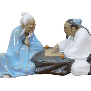 May include: Ceramic figurine depicting two seated figures playing a board game. One figure wears a light blue robe, the other a white robe. The game board is dark gray, and the pieces are orange.