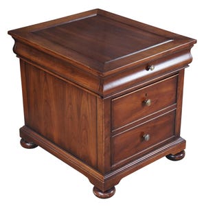 May include: A dark brown wooden end table featuring a flat top surface, one shallow top drawer, and two deeper lower drawers. Each drawer is equipped with a small, round, metallic knob. The piece is supported by four rounded, bun-style feet.