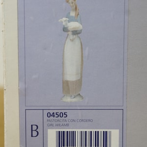 May include: A Lladro porcelain figurine of a woman holding a lamb. The woman wears a blue top and a long white apron. The figurine is on a light blue background. The box has the text "04505 PASTORCITA CON CORDERO GIRL WILAMB".