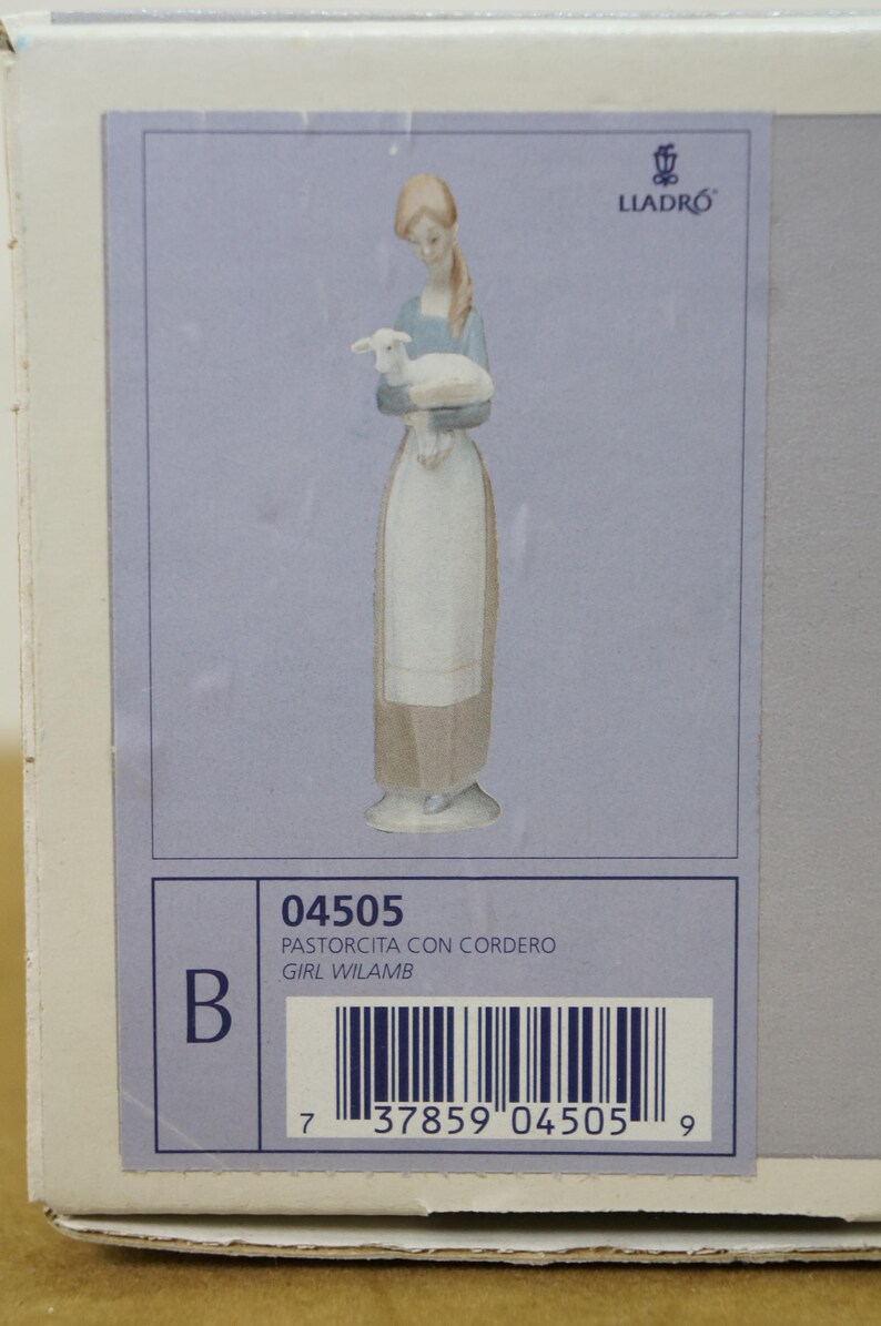 May include: A Lladro porcelain figurine of a woman holding a lamb. The woman wears a blue top and a long white apron. The figurine is on a light blue background. The box has the text "04505 PASTORCITA CON CORDERO GIRL WILAMB".