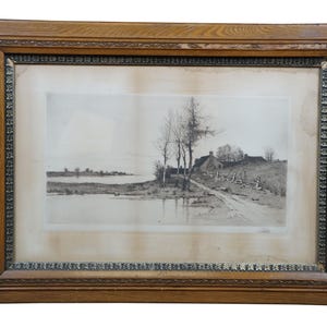 May include: A framed black and white landscape print. The artwork depicts a rural scene with a body of water, trees, a dirt road, and a small building. The frame is made of wood with a decorative border.